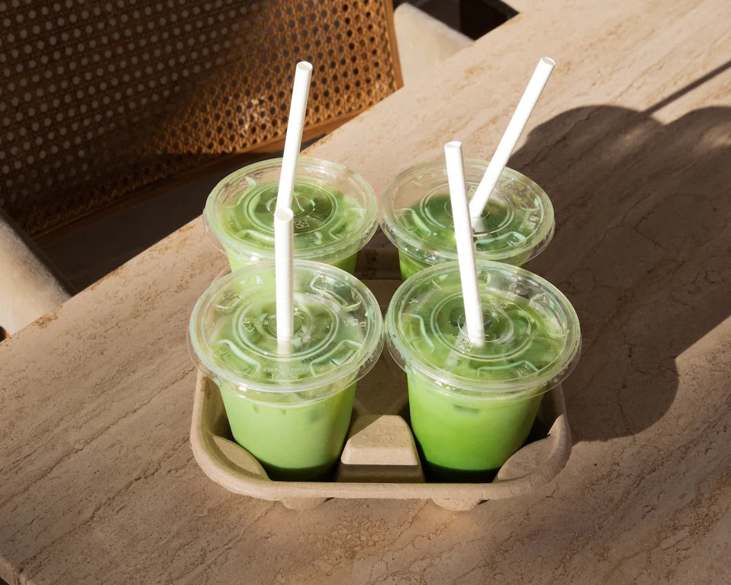 Four green mathca drinks in clear plastic cups with straws on a wooden table.